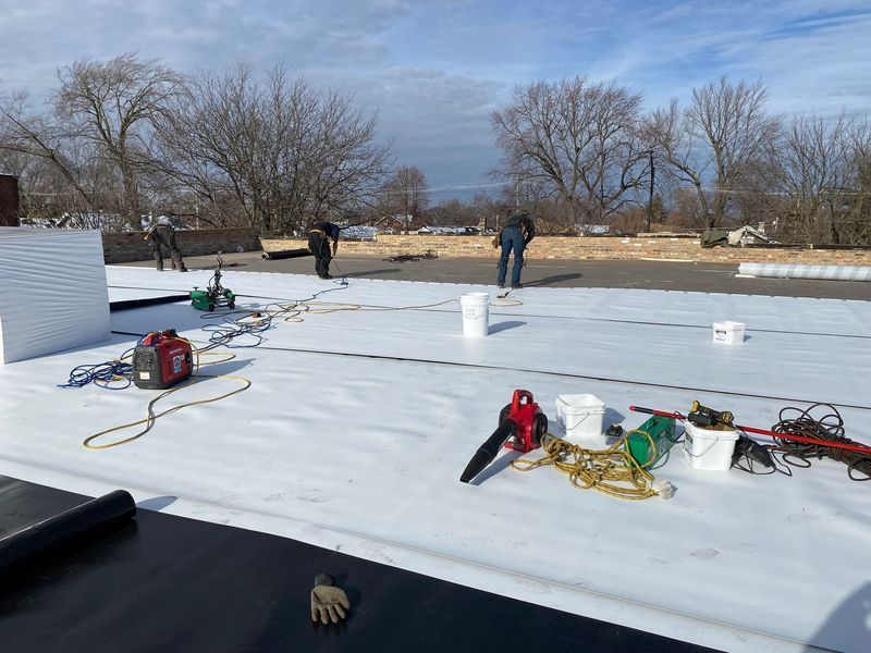 Workers installing a white roof, using tools and materials on a flat rooftop on a sunny day.
