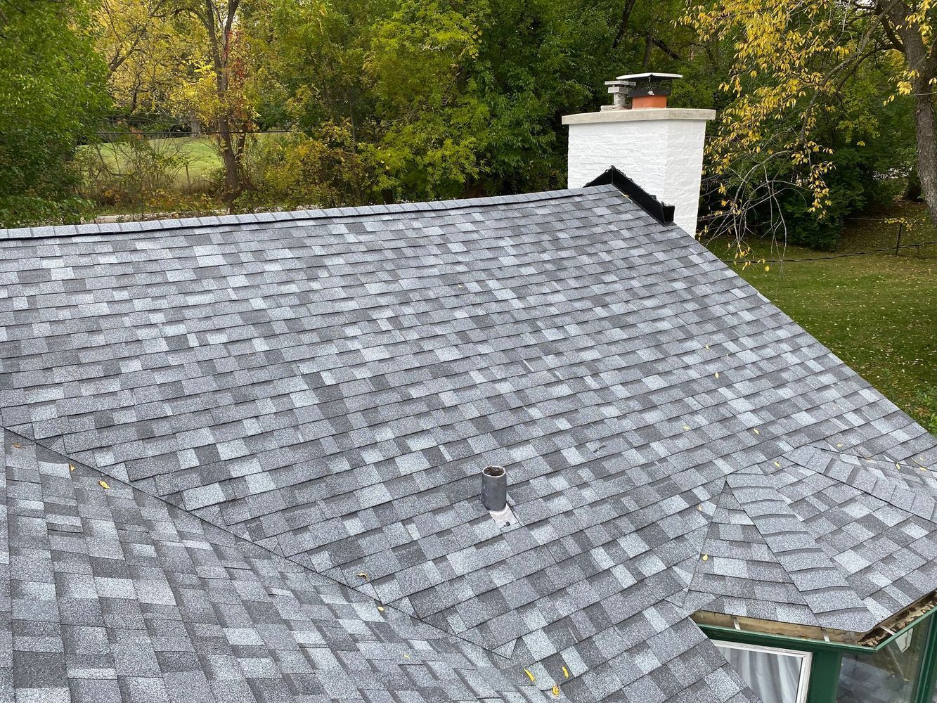 Gray shingle roof on a house with a white chimney; green trees and grass in background.