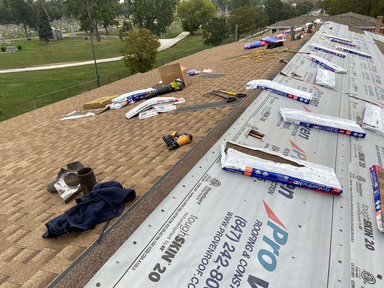 Roof with materials and tools; workers installing shingles. Green grass and trees in background.