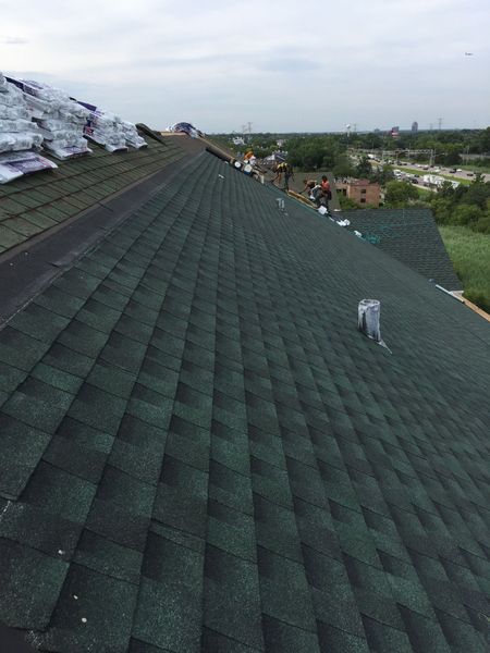 Green shingle roof being installed by workers. Overcast sky and green landscape in the background.