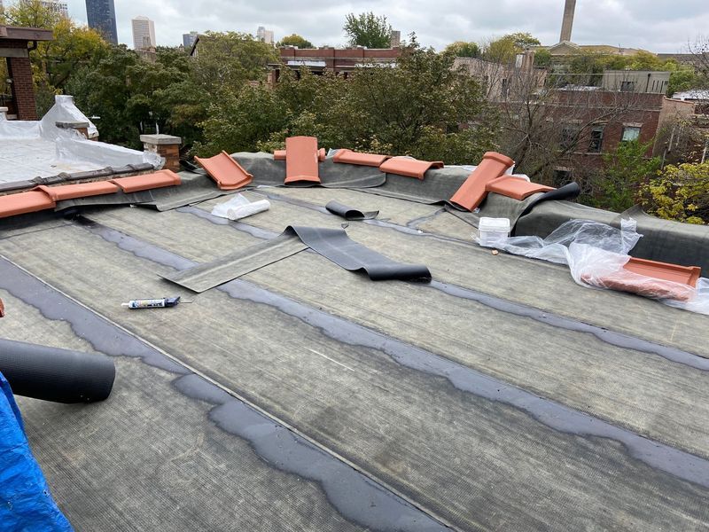 Rooftop under construction, featuring tiles, sealant, and rolled roofing materials. Buildings and trees in the background.