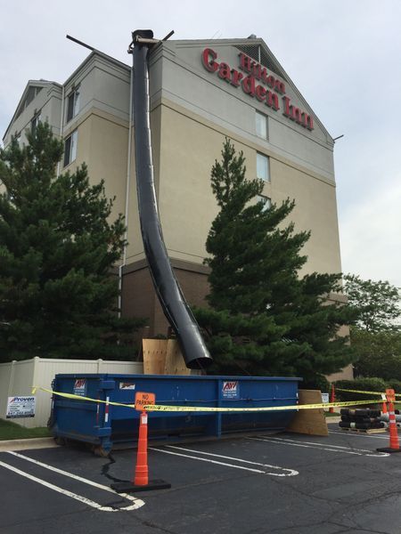 A black debris chute extends from a Hilton Garden Inn to a blue dumpster in a parking lot.