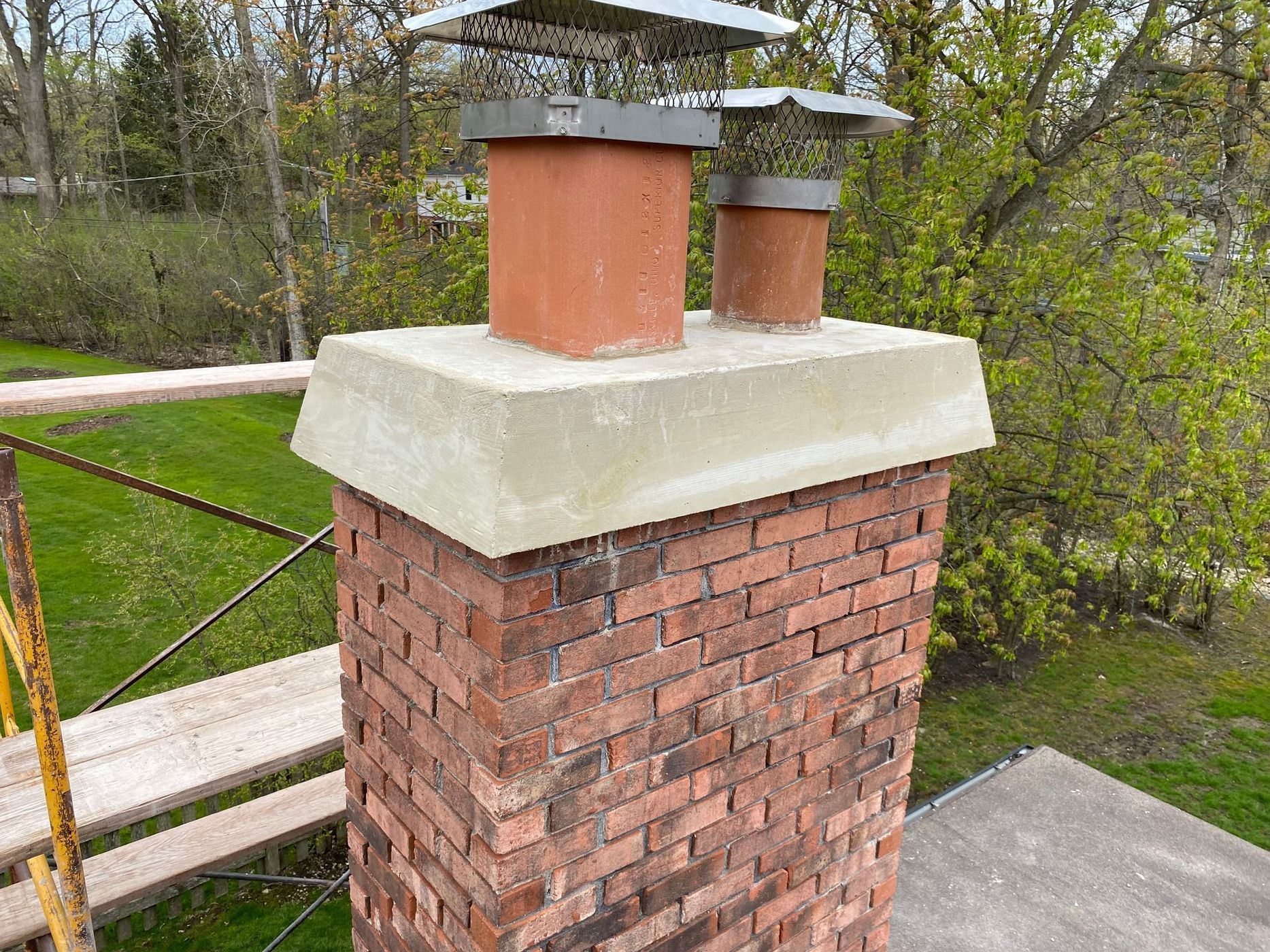 Brick chimney with concrete cap and two flue pipes topped with metal covers.