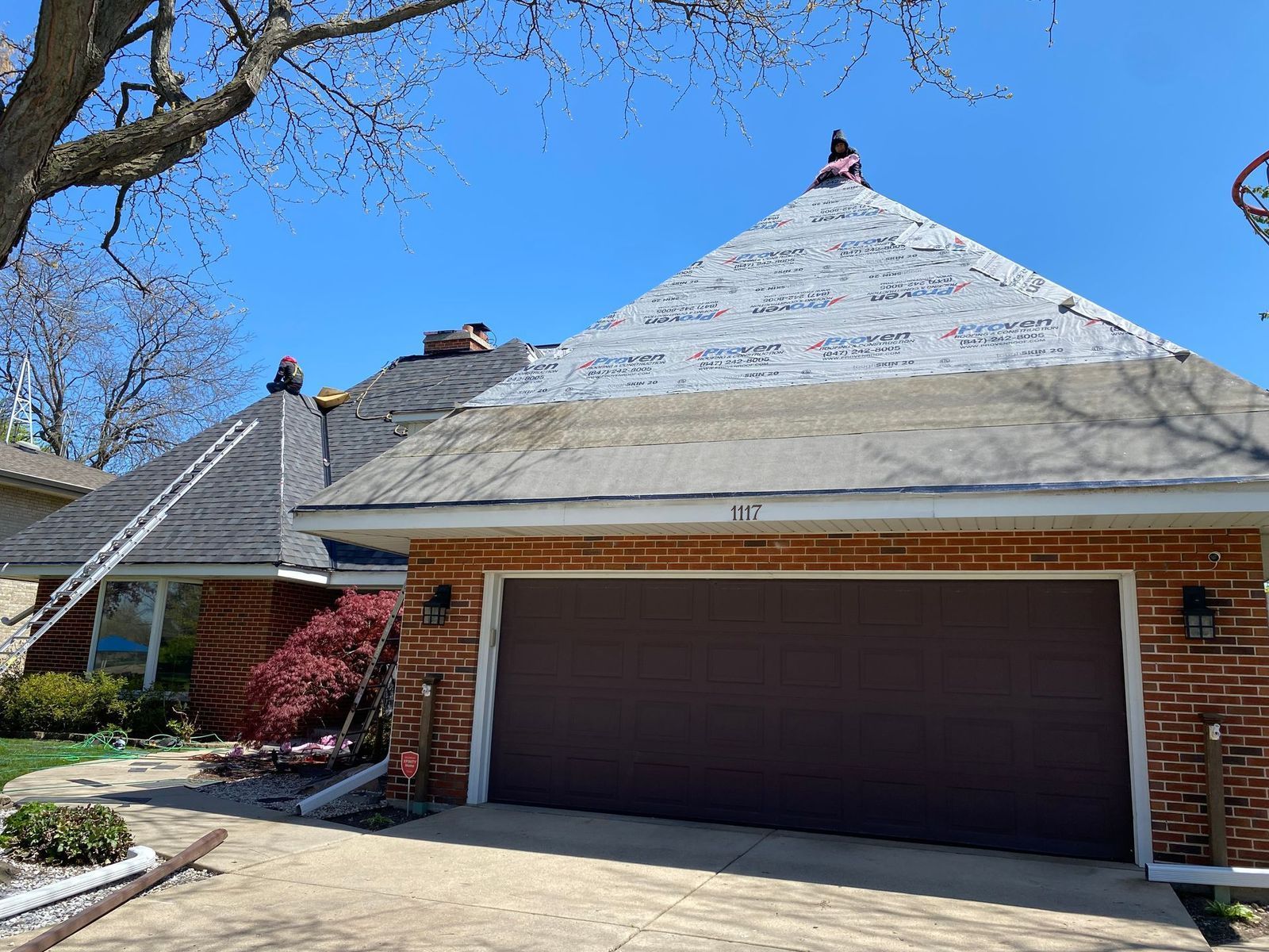 Roofers on a house with a partially replaced roof; red brick exterior, brown garage door, and blue sky.