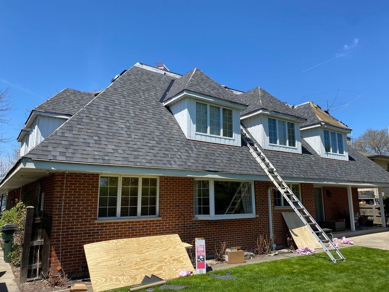 House with new gray roof, brick exterior, blue sky; ladder and plywood lean against the building.