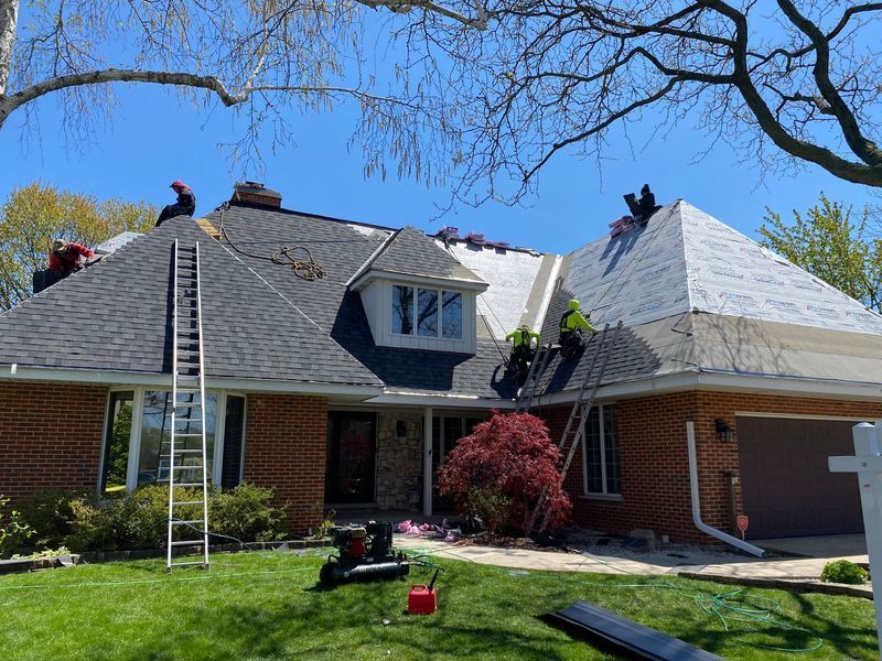 Roofers replacing shingles on a brick house with multiple roof sections; sunny day.