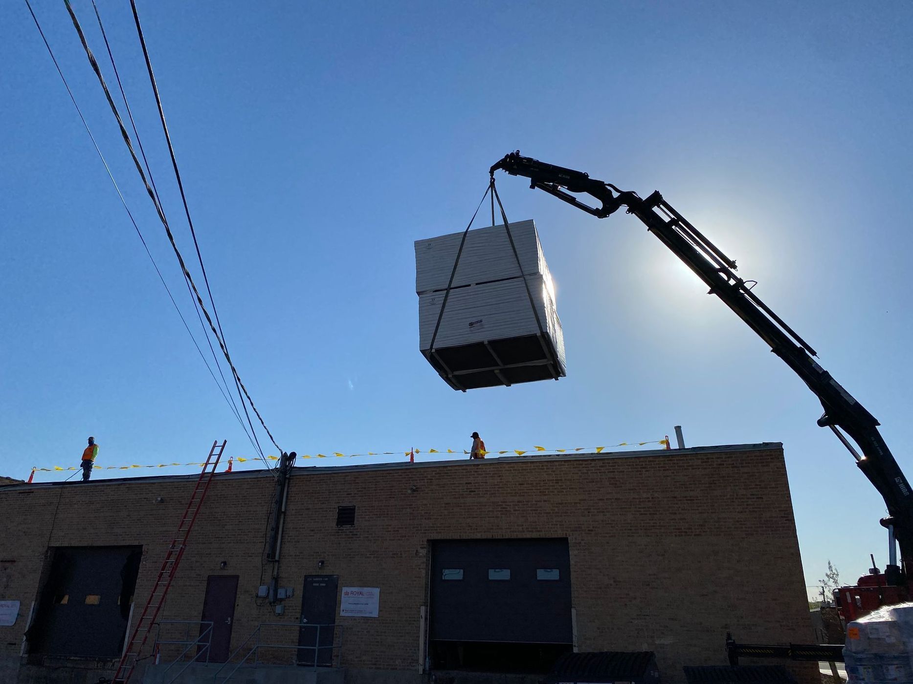 A crane lifts a large, boxed unit above a brick building under a bright blue sky.