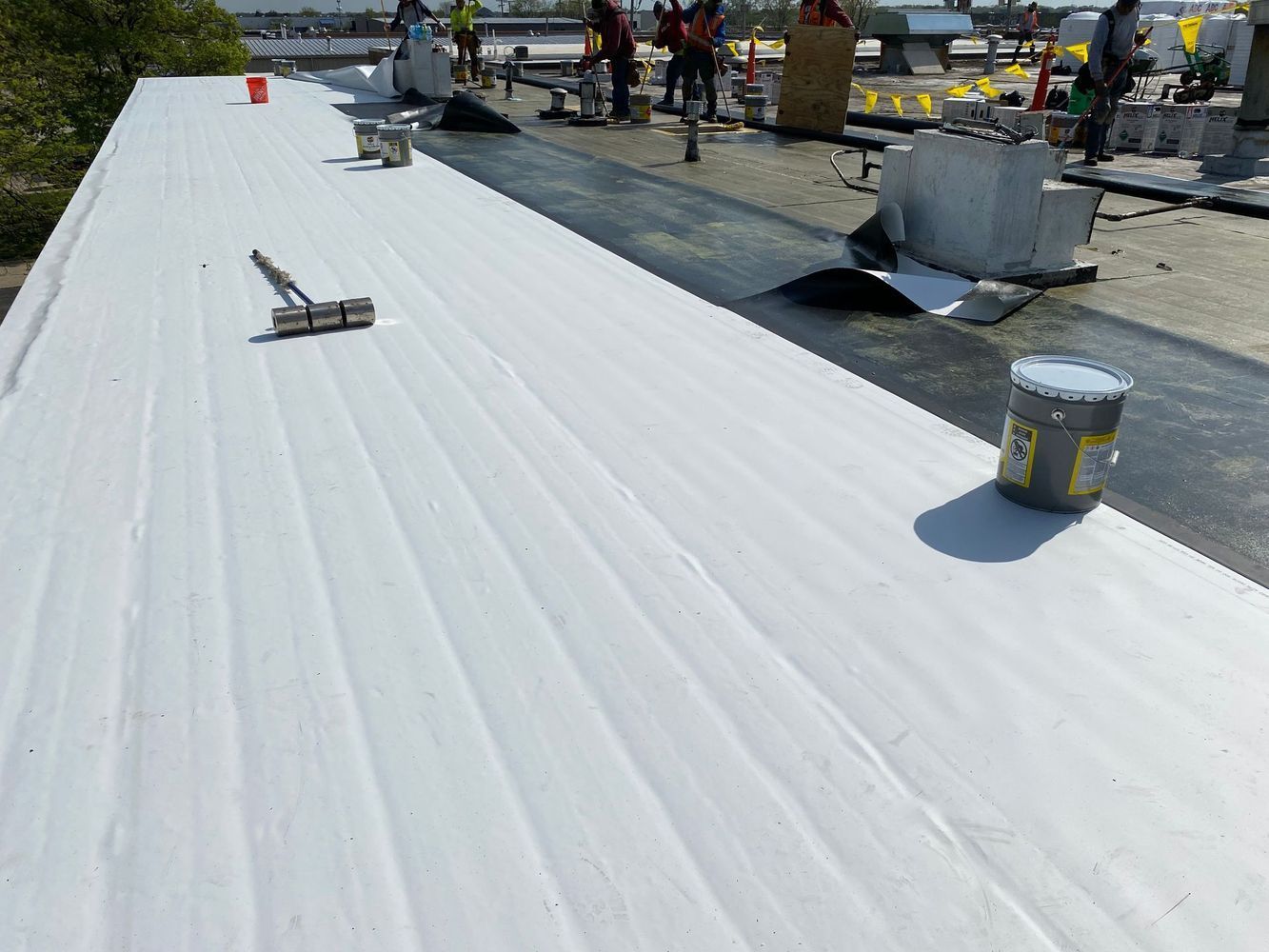 Workers applying white coating to a flat roof. Paint cans and tools visible, sunlight overhead.
