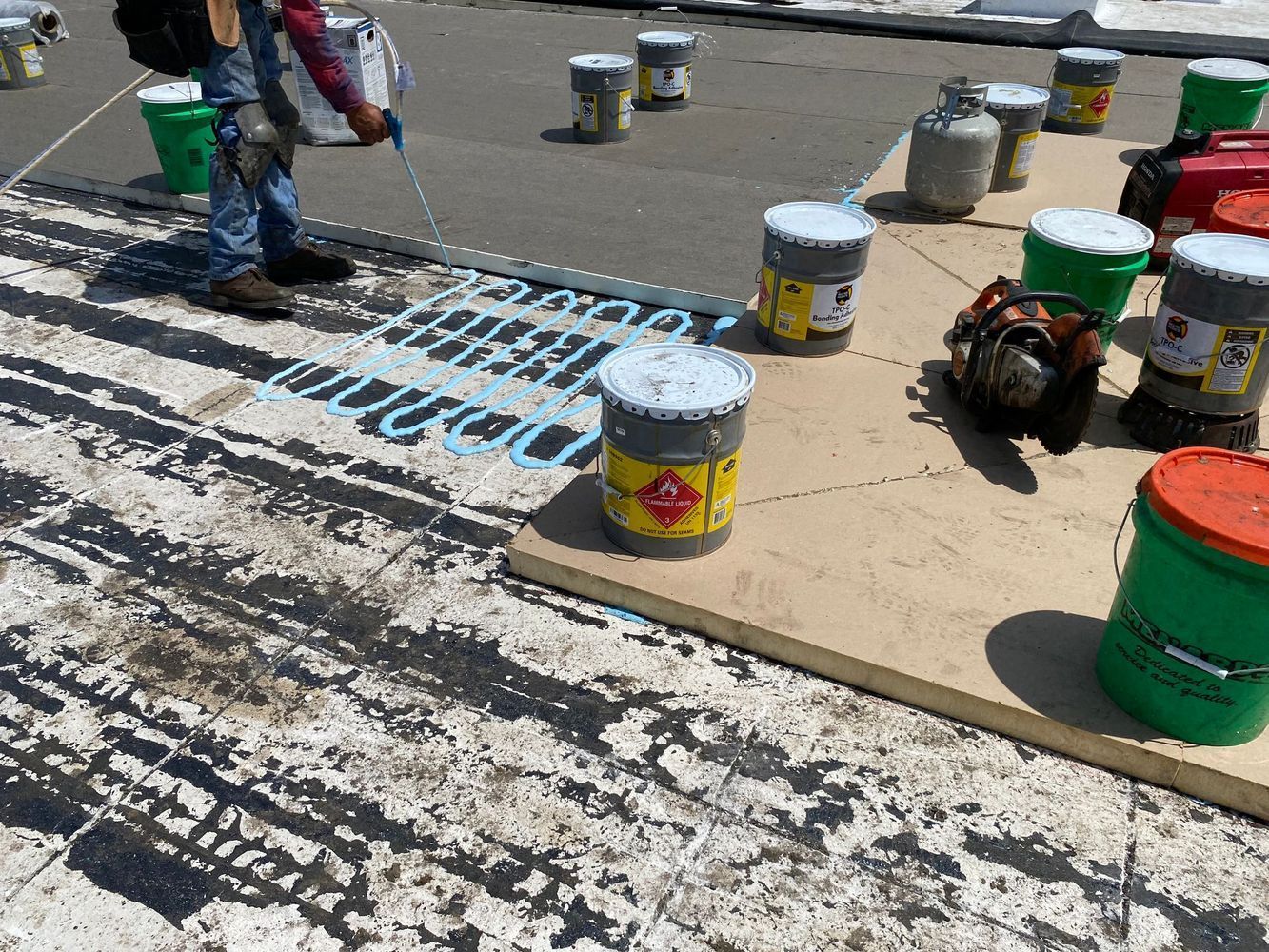 Roofer applying sealant on a flat roof, surrounded by tools and buckets of materials.