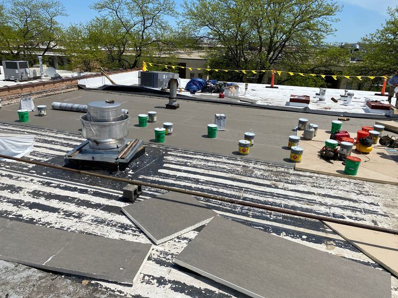 Rooftop undergoing repairs; gray roofing material, tools, equipment, and paint cans are visible, with a sunny, outdoor setting.