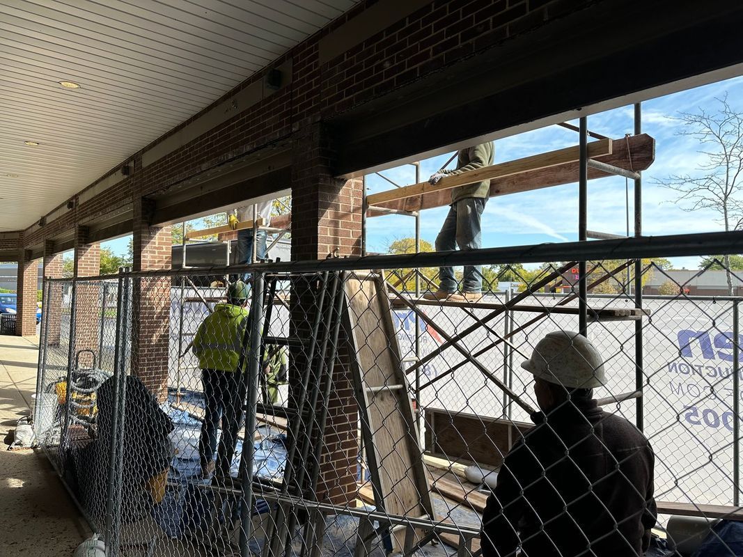 Construction workers building near a brick building. One worker is on scaffolding, others on ground. Chain-link fence present.