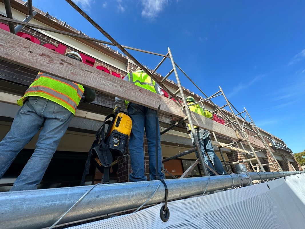 Construction workers on scaffolding, installing boards on a building. They wear safety vests against a blue sky.