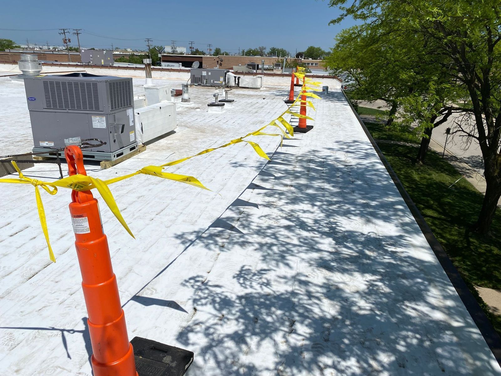 Rooftop with orange safety cones and yellow caution tape near HVAC units and trees.