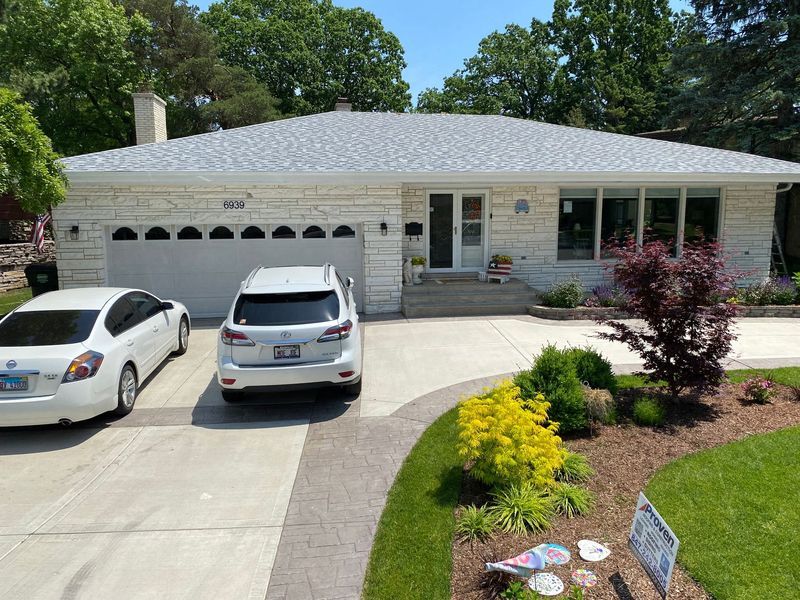 White suburban home with attached garage, cars in driveway, landscaped yard on sunny day.