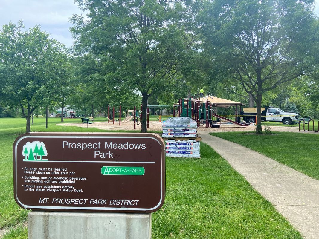 Prospect Meadows Park sign at playground with construction. Gray walkway, green trees, cloudy sky.
