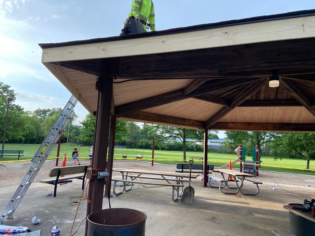 Person in a yellow vest repairing a wooden pavilion roof in a park.