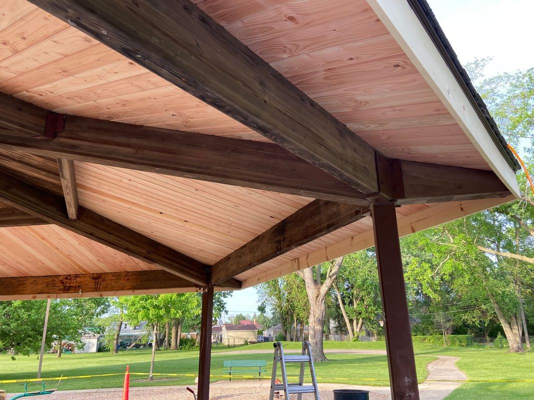 Wooden gazebo with a red-brown roof and dark brown support beams, outdoors with trees and grass.