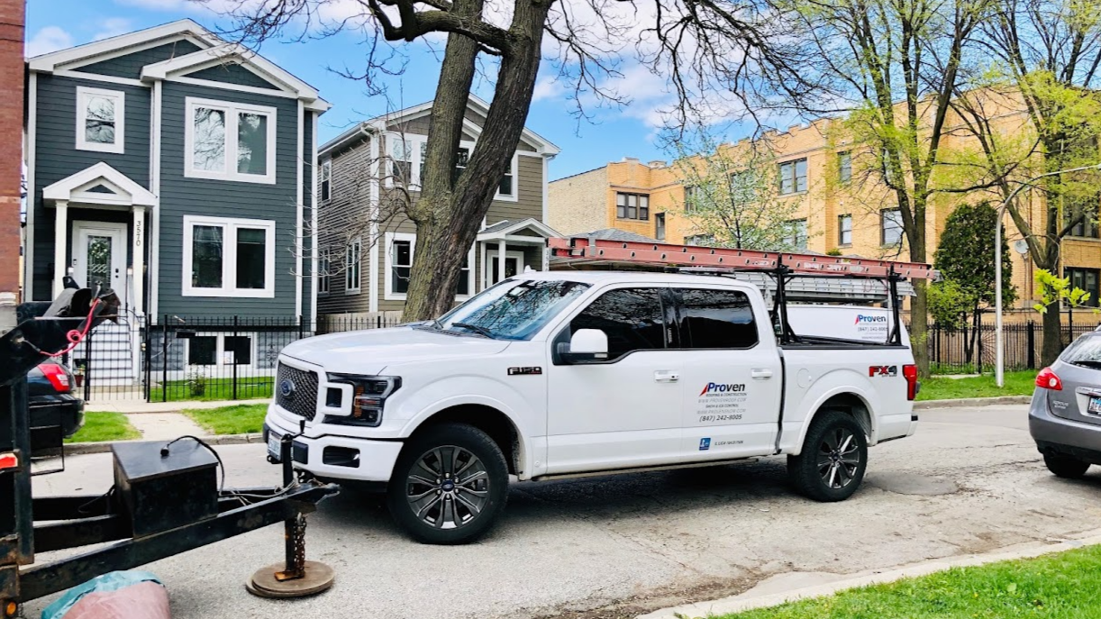 White pickup truck with ladder and trailer parked on a street in front of houses on a sunny day.