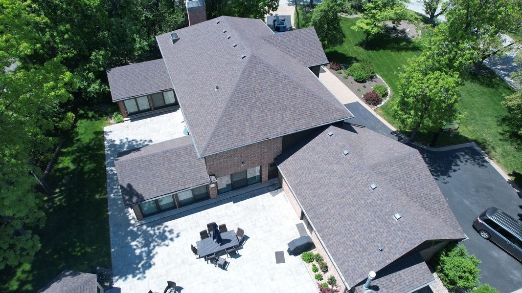 Aerial view of a brown-roofed house with a patio and trees on a sunny day.