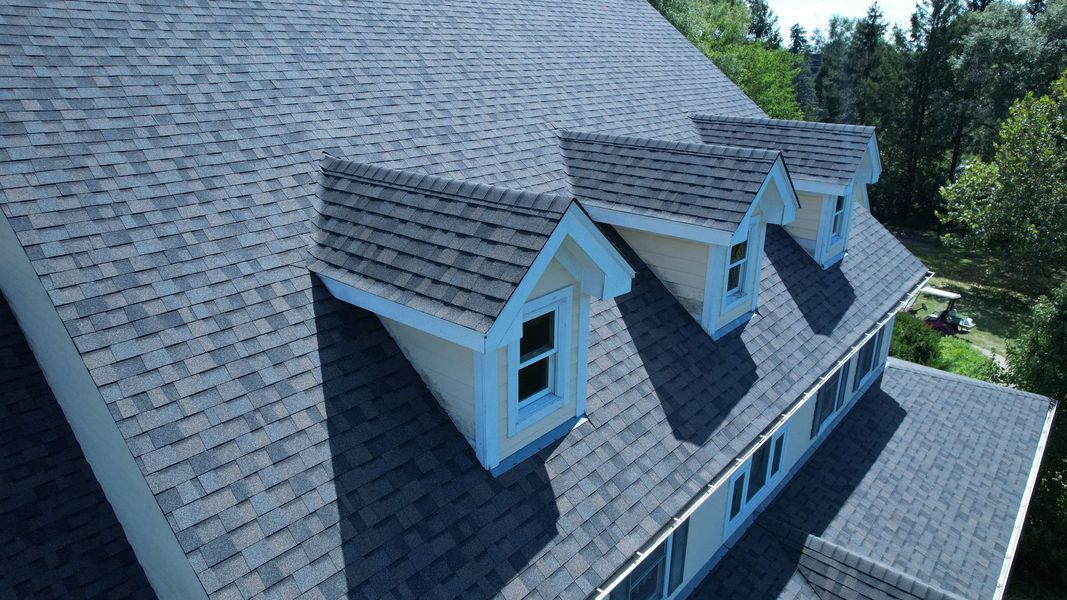 A roof of a house with three dormers, blue shingles, yellow walls, and white trim.
