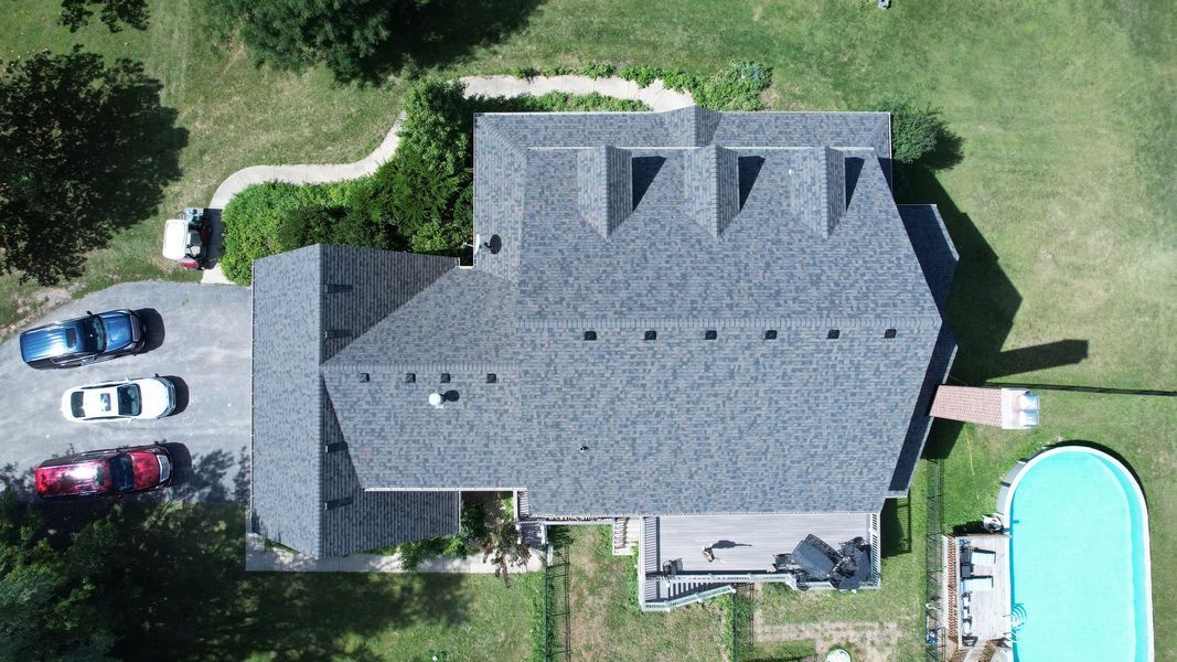 Aerial view of a gray-roofed house with cars parked in the driveway and a swimming pool in the yard.