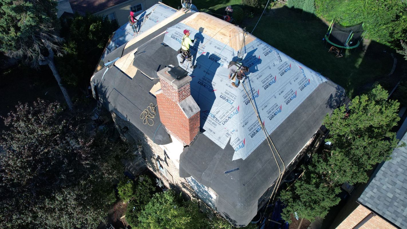 Workers repairing a roof with new shingles. They stand near a brick chimney, surrounded by trees.