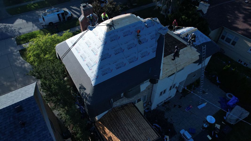 Aerial view of roof repair on a two-story house. Workers install shingles on a partially completed roof.