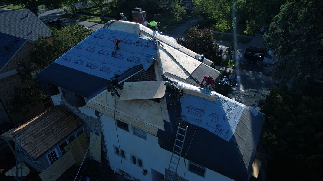Workers repairing a roof. One person on the roof. Bright blue sky, houses, and trees.