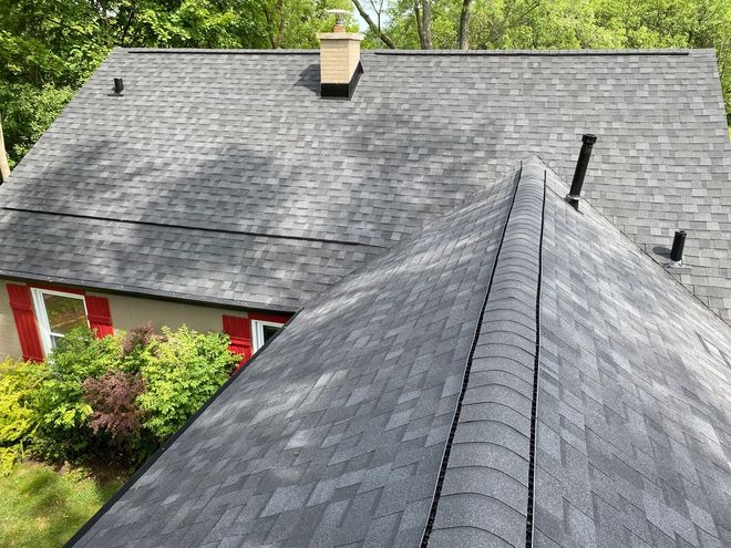 Dark gray shingled roof with a central peak, chimney, and pipes against a backdrop of trees.