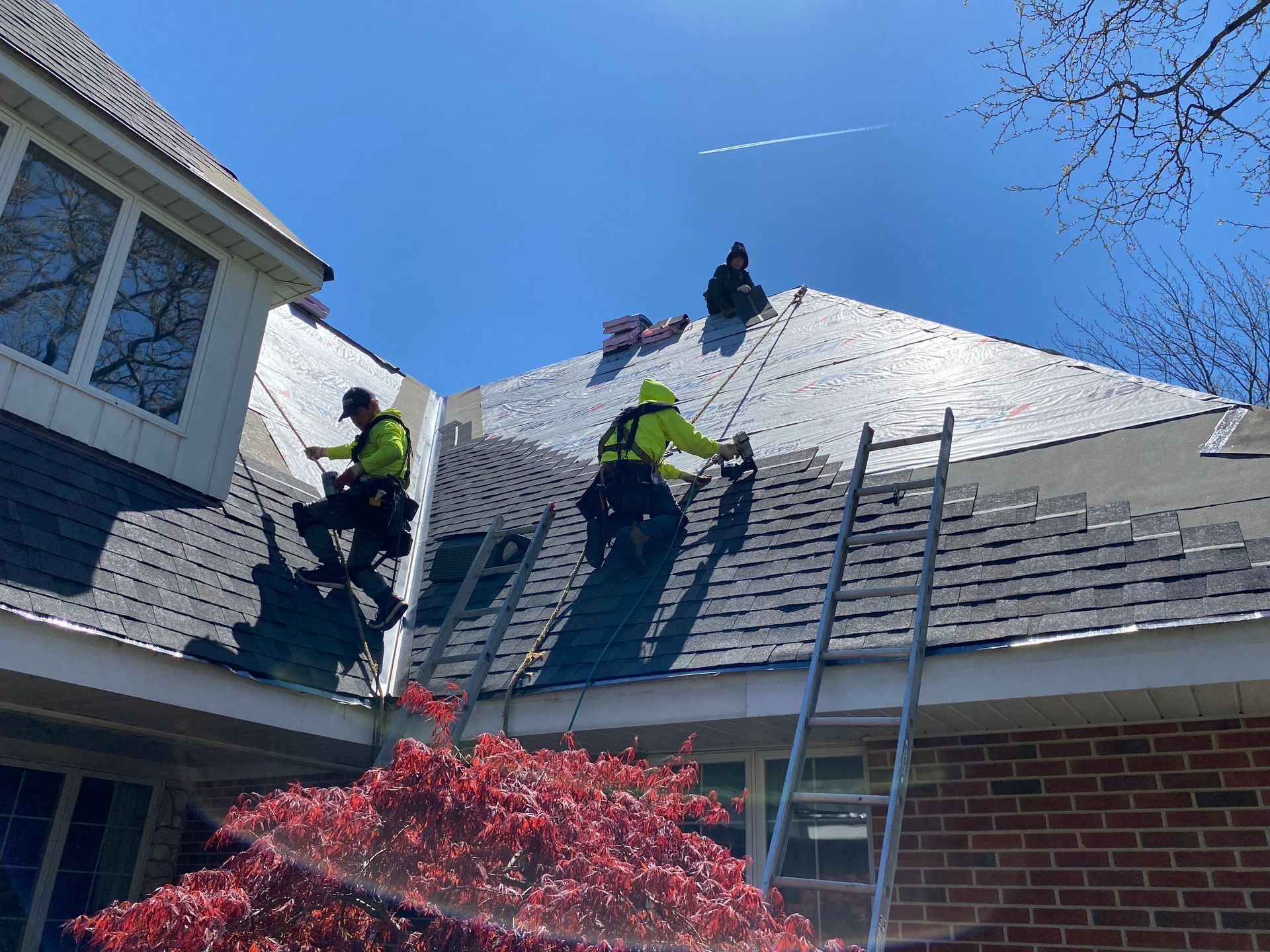 Workers on a roof with ladders, wearing safety gear under a blue sky, repairing shingles.