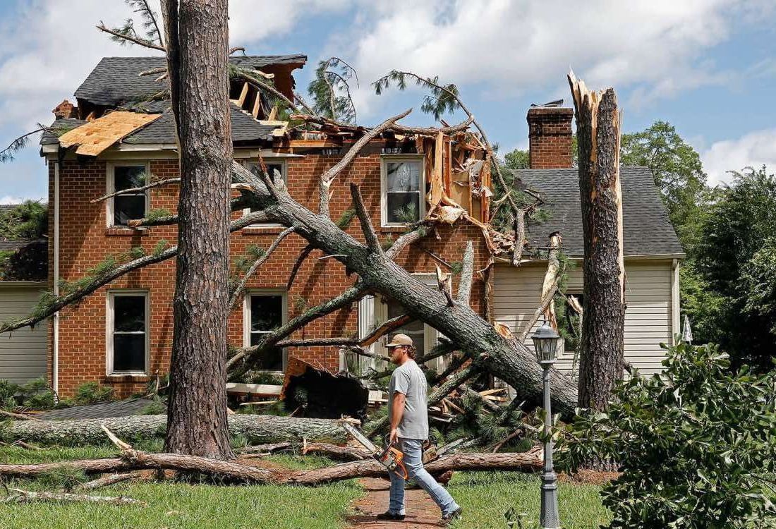 Man walks past a damaged brick house; trees fallen on the roof after a storm.