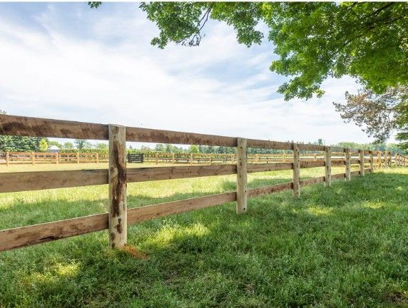 Wooden split-rail fence beside a grassy field under green trees and a blue sky