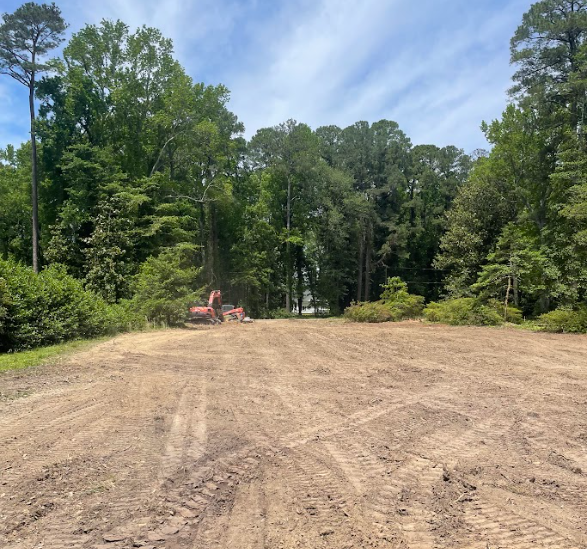 Cleared dirt lot bordered by tall green trees, with a small red excavator in the distance under a blue sky