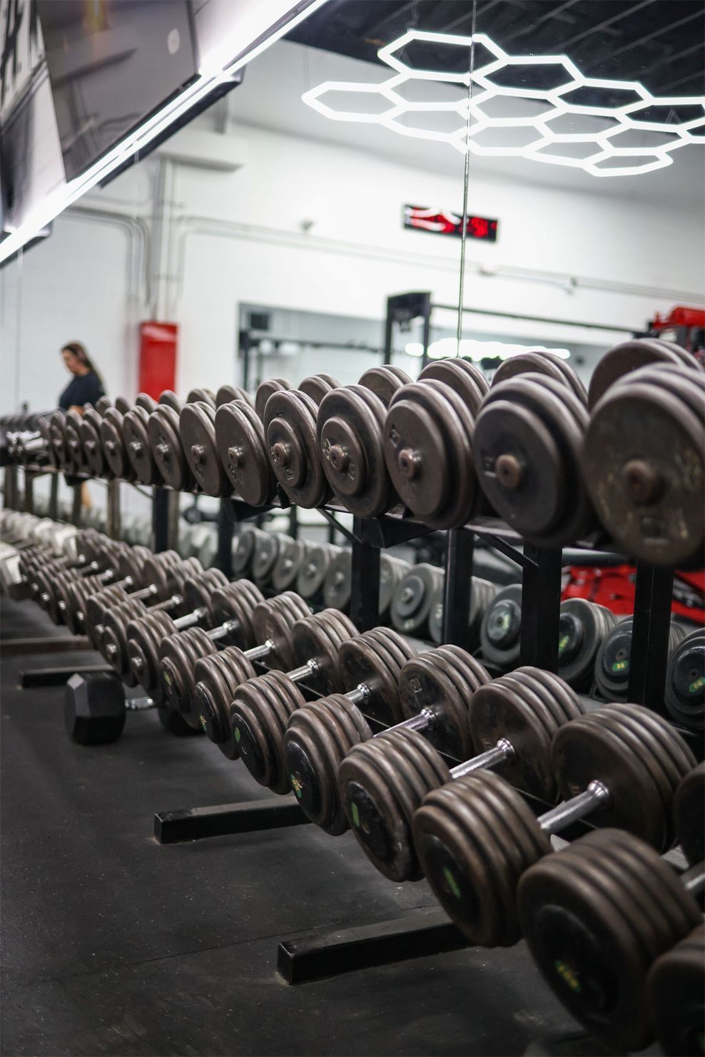 Rows of dumbbells in a gym setting, with a person visible in the background.