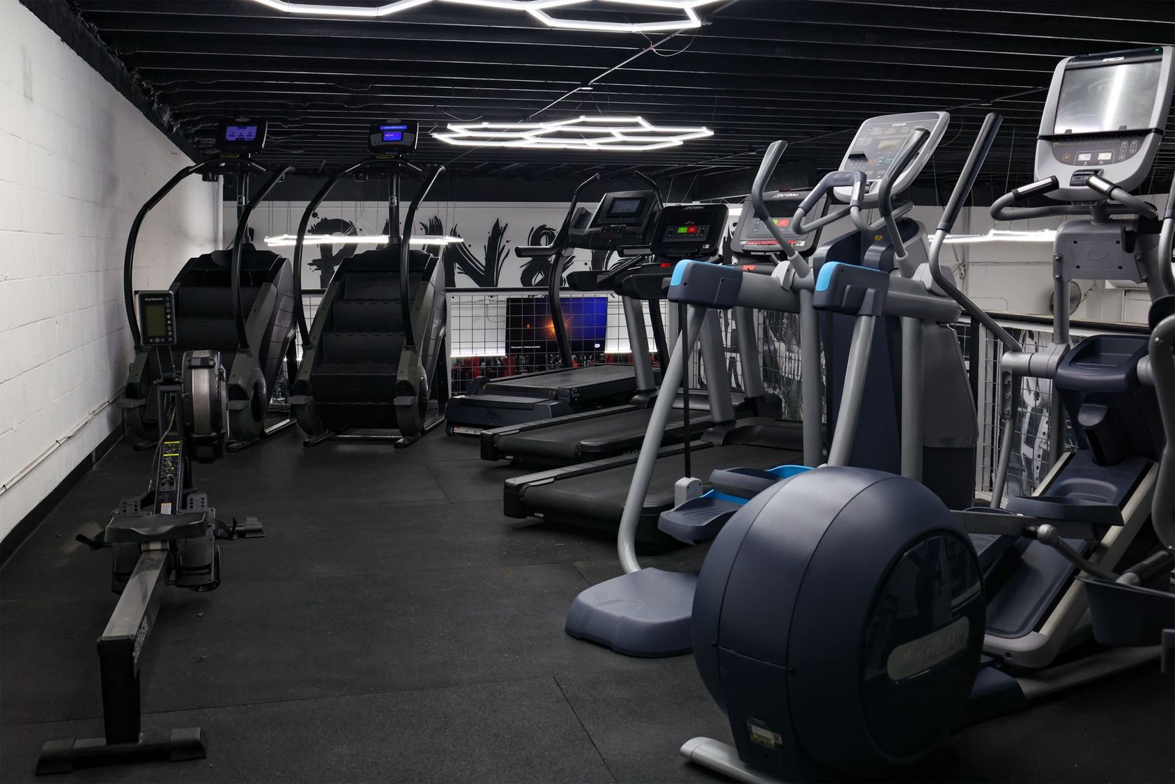 A gym interior with exercise machines including elliptical trainers, stair climbers, treadmills, and a rowing machine.
