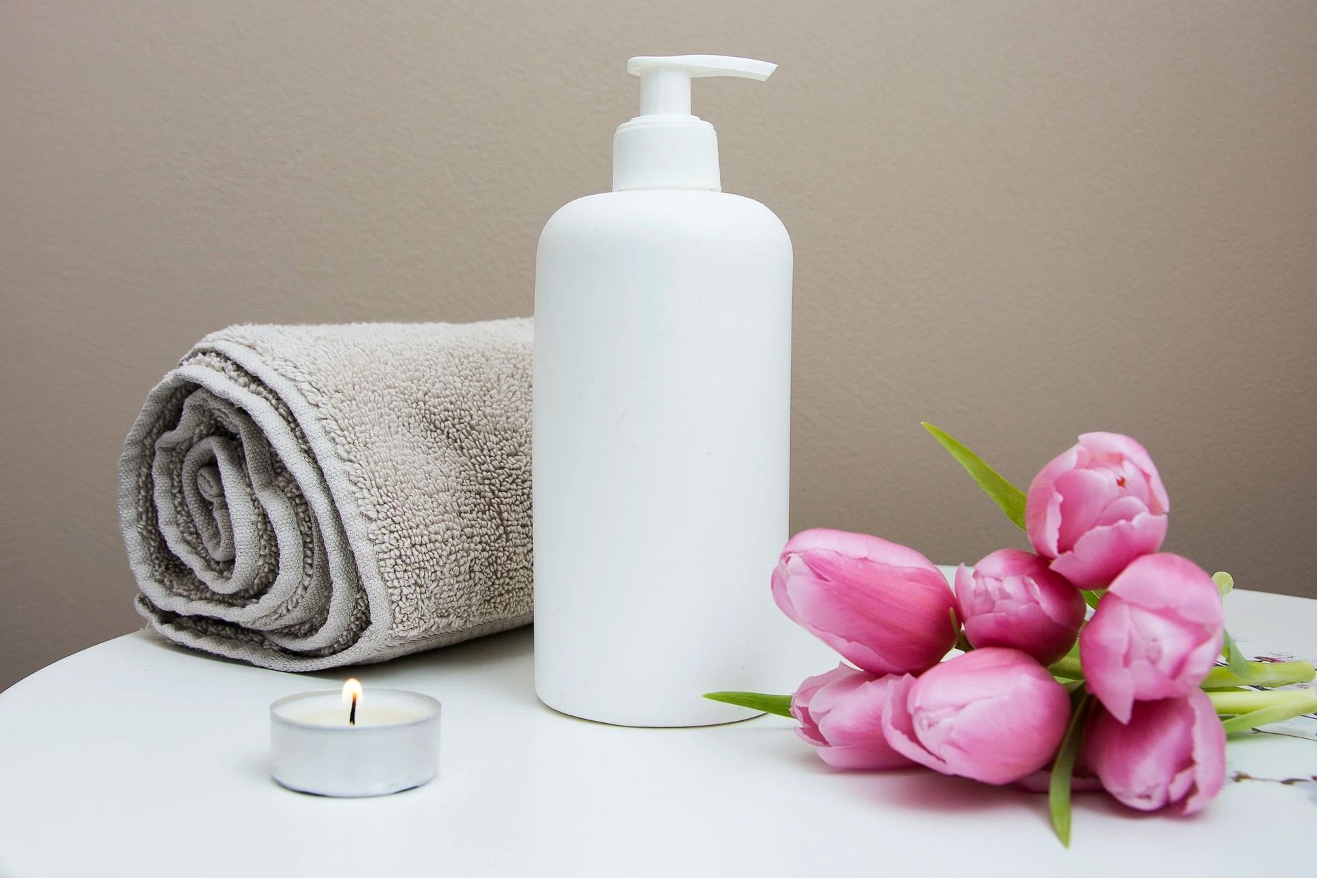 White lotion bottle, rolled towel, lit candle, and pink tulips on a white surface.