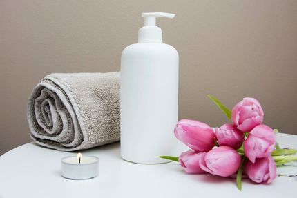 White lotion bottle, rolled towel, lit candle, and pink tulips on a white surface.
