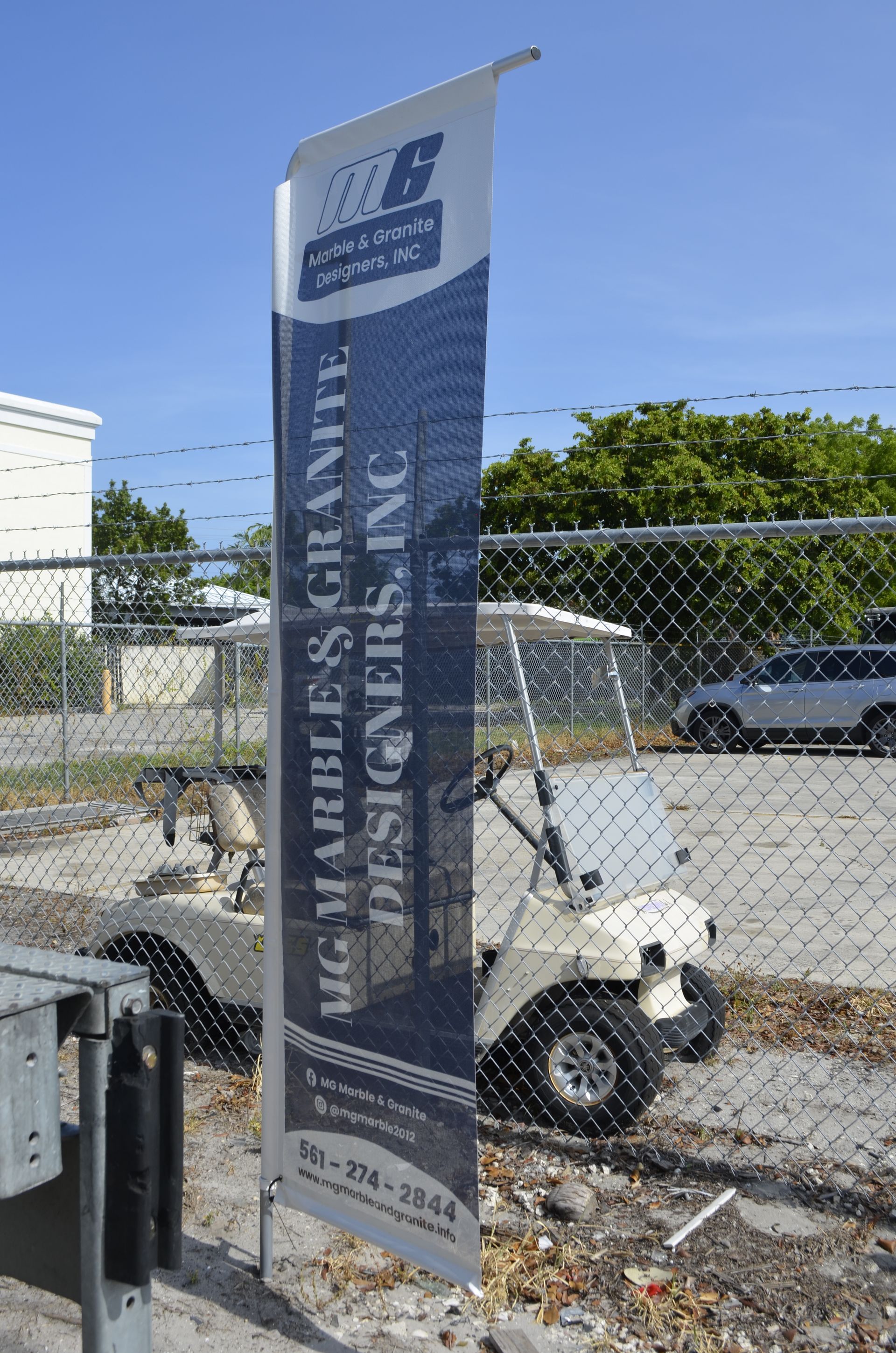 Flag sign for MG Marbles & Granite Designers, Inc. by a golf cart and fence.