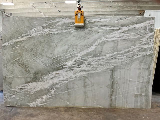 Large slab of pale green marble with white veining, being lifted by a yellow crane in a warehouse.