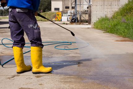 Worker in yellow boots pressure-washing a concrete surface with a hose outdoors.