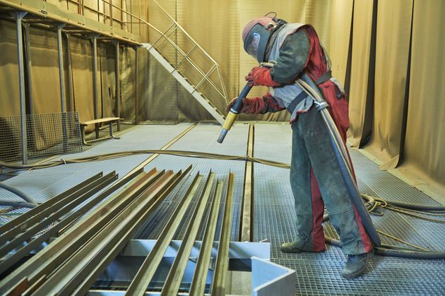 Worker in protective suit sandblasting metal beams inside an industrial facility.