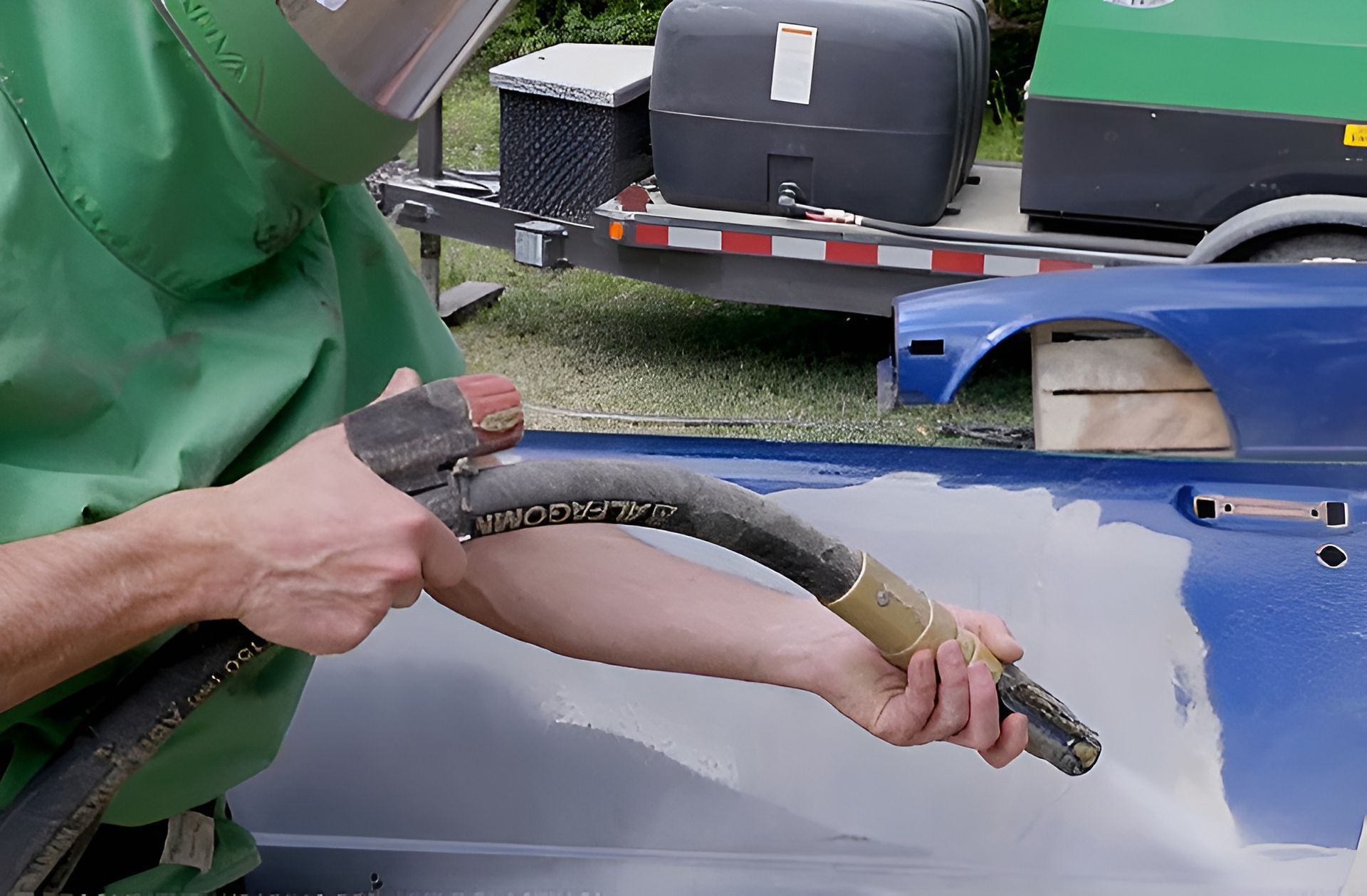 A worker in a protective green suit uses a media blaster to strip paint from a blue car door panel outdoors.