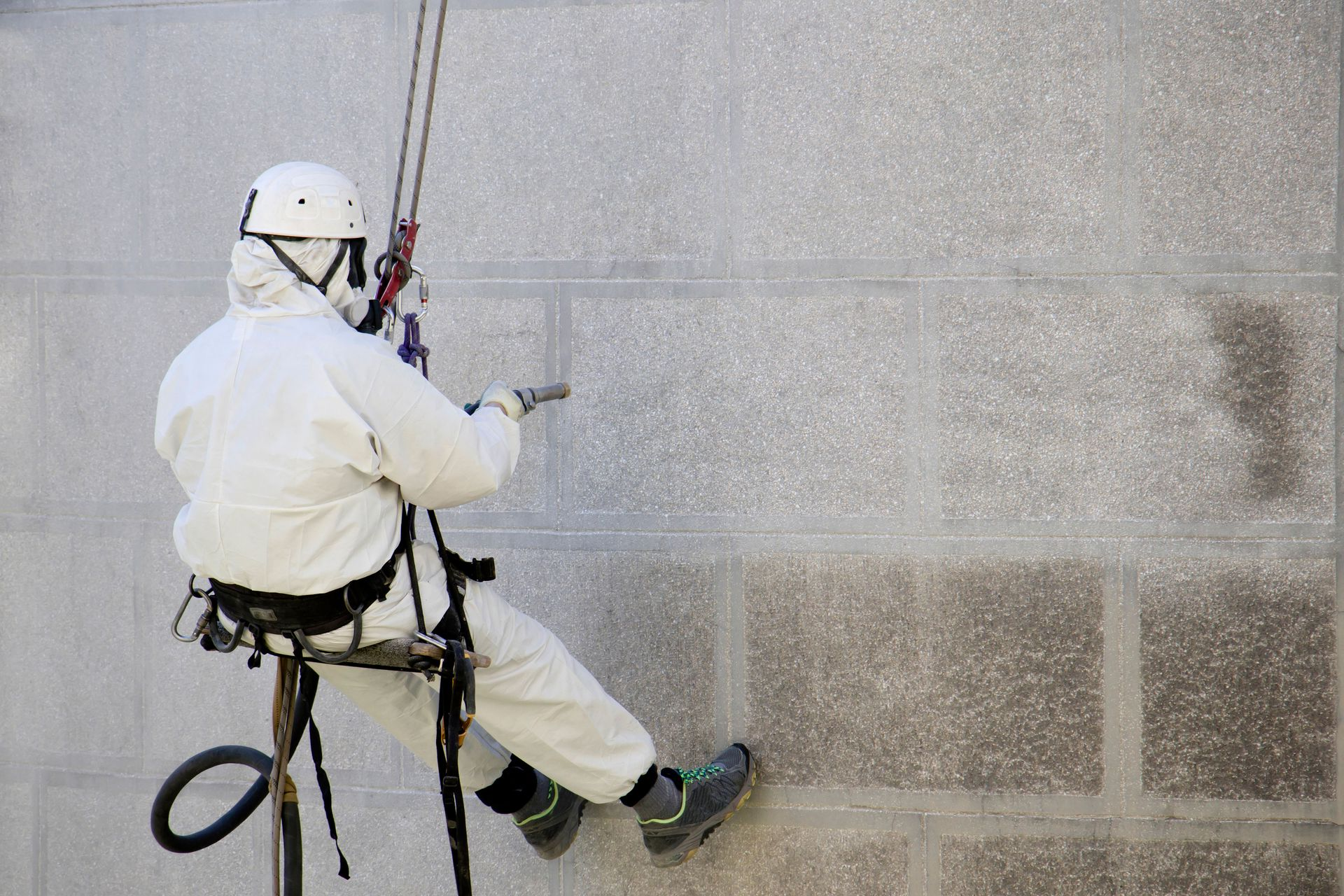 Worker in protective suit suspended by ropes, spraying a wall.