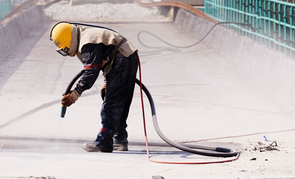 Worker sandblasting a bridge, wearing protective gear and helmet.
