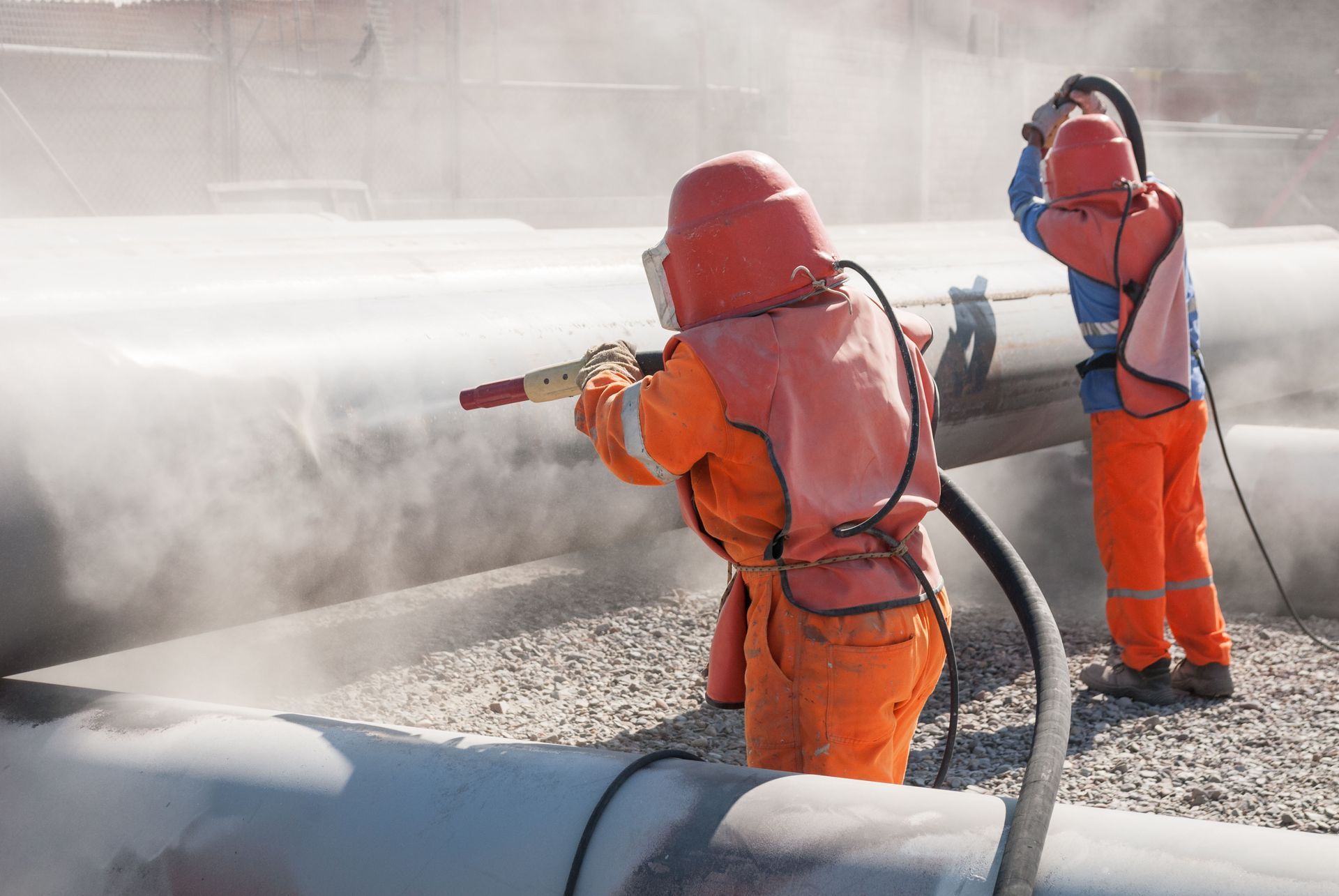 Two workers in safety suits sandblasting pipes, creating a cloud of dust outdoors.