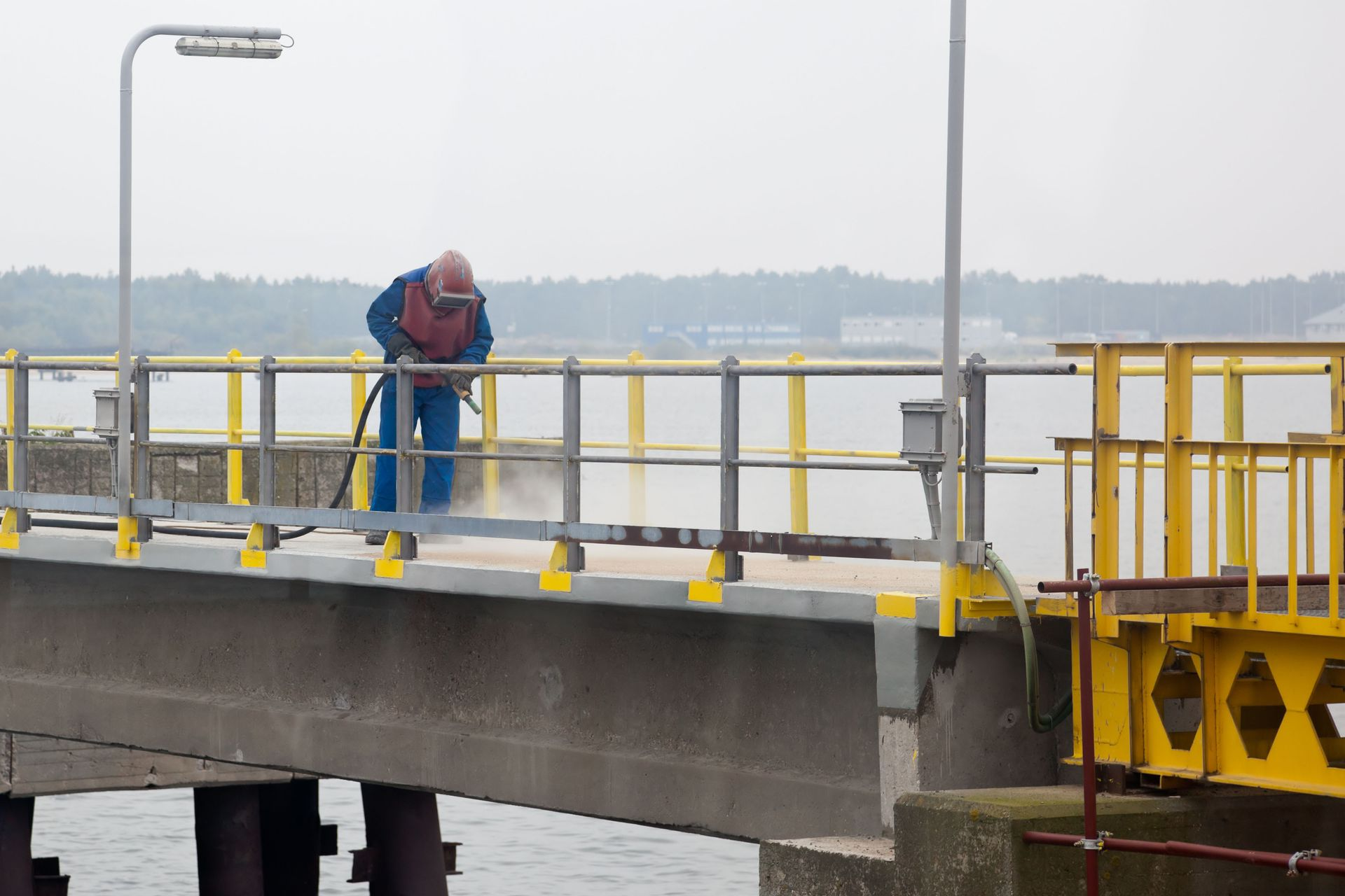 Man in protective gear sandblasting a pier with yellow railings, cloudy sky over water.