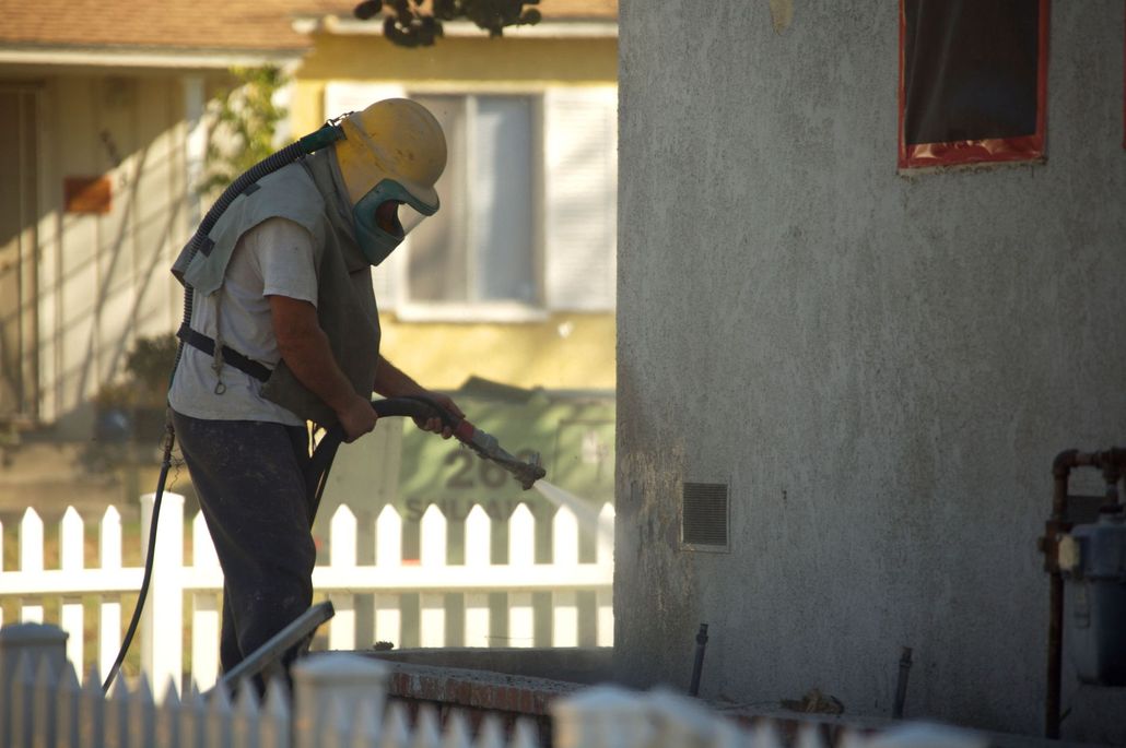 Person in safety gear sandblasting a building's exterior near a white picket fence.