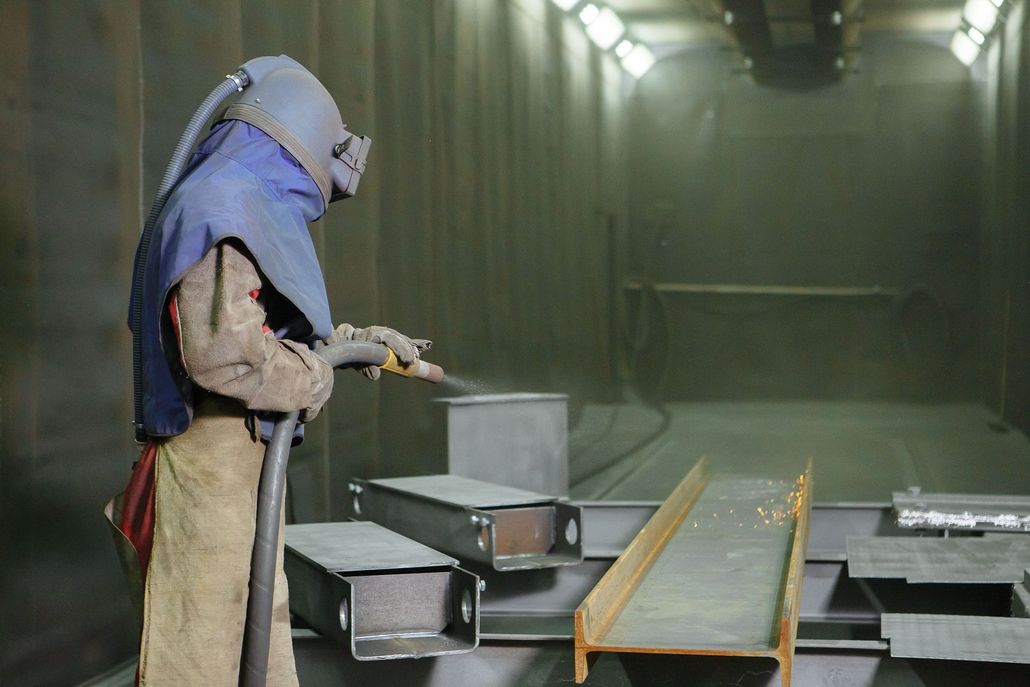 Person in protective gear sandblasting metal beams inside an industrial workshop.