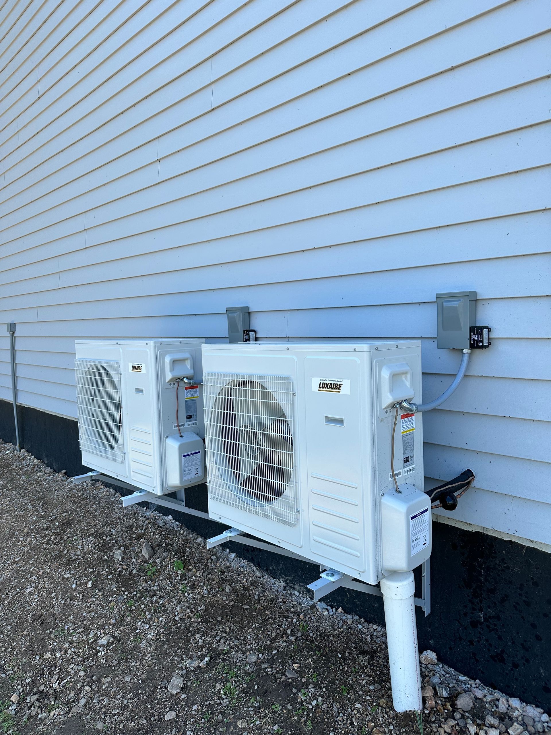 Two white heat pump units mounted on the side of a building with electrical boxes and pipes.