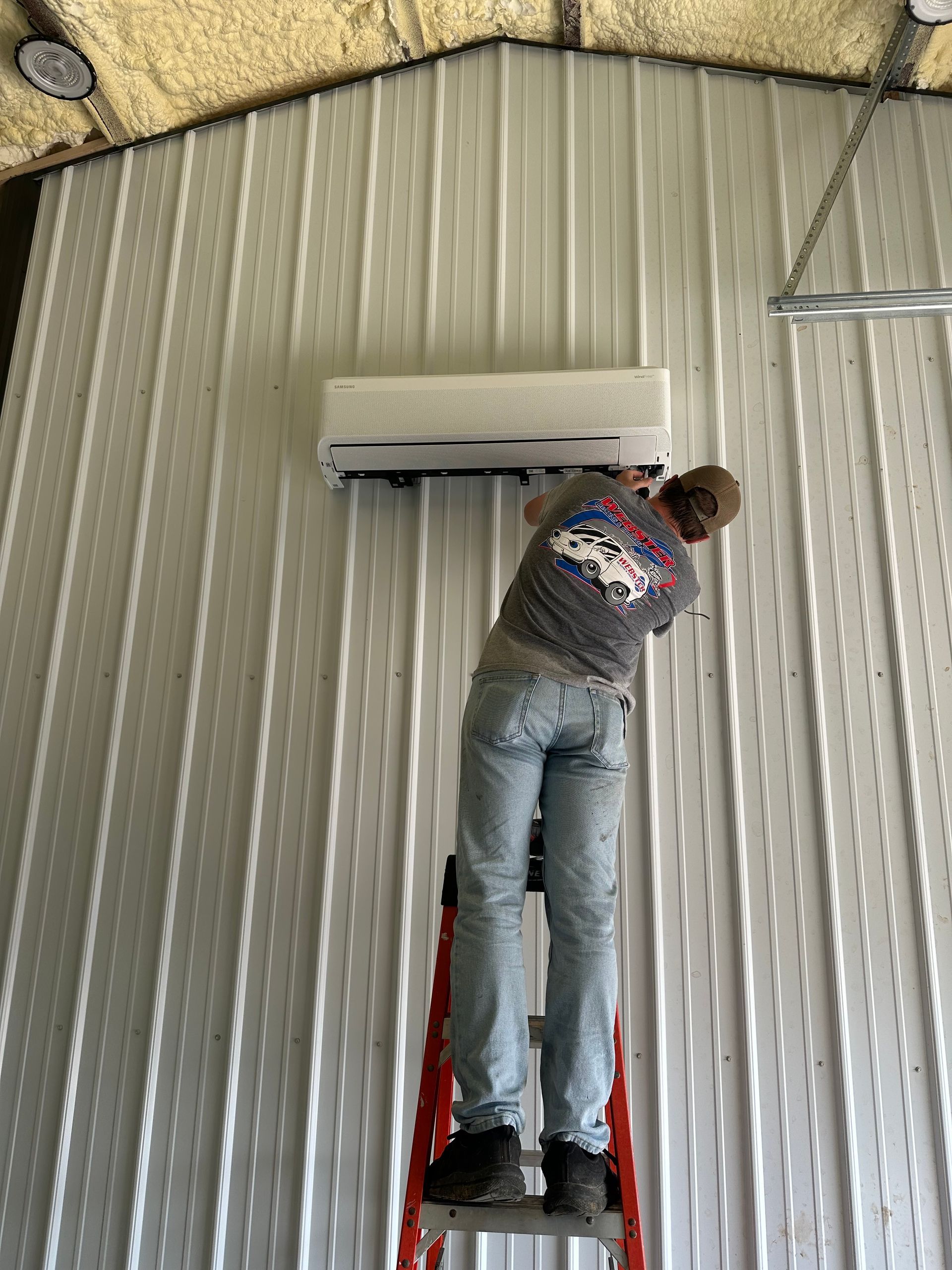 Person on a ladder installing an air conditioning unit on a white metal wall inside a building.